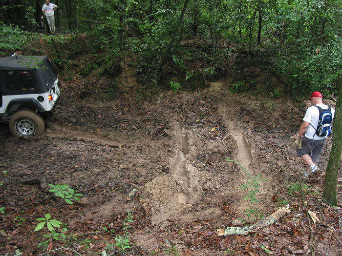 White Jeep Wrangler stuck in mud on a wooded trail. A man in a red hat walks away from it