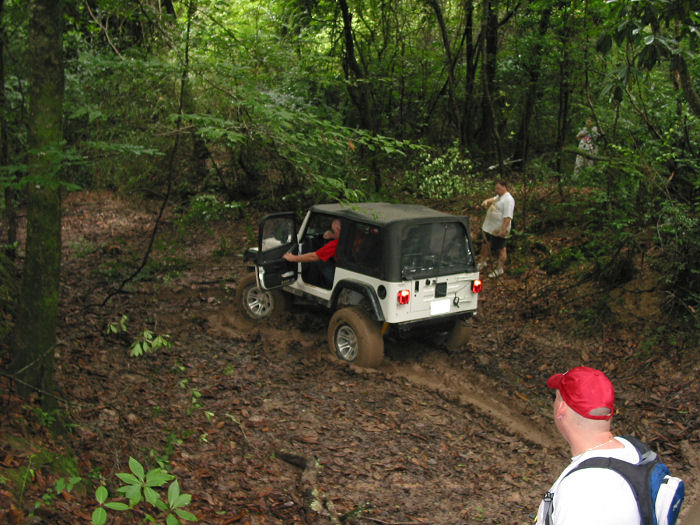 White Jeep Wrangler with black soft top driving on a muddy trail in a forest