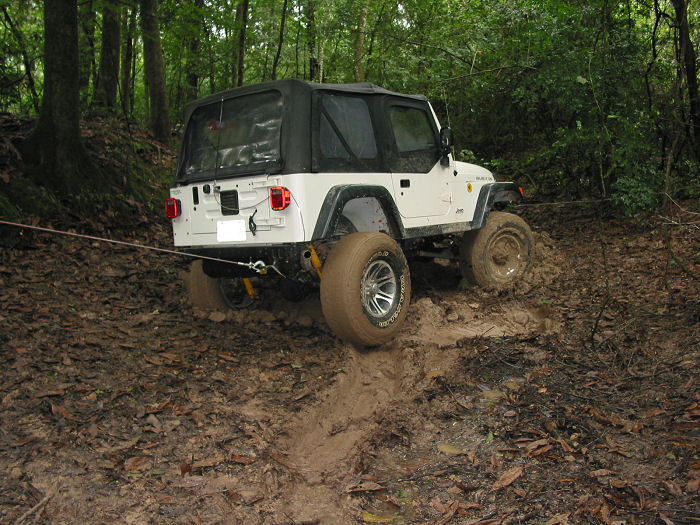 White Jeep Wrangler with soft top driving through a muddy trail in the woods. A tow rope is attached to the rear