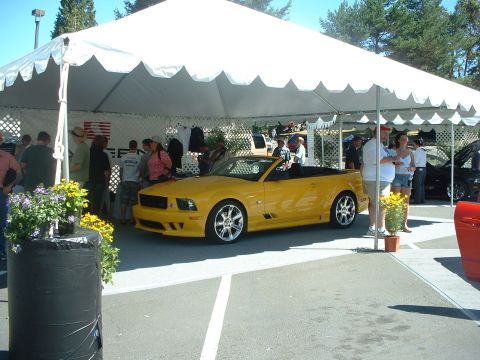 1999 Ford Crown Victoria - A yellow convertible Mustang is displayed under a tent at a car show, with spectators around.