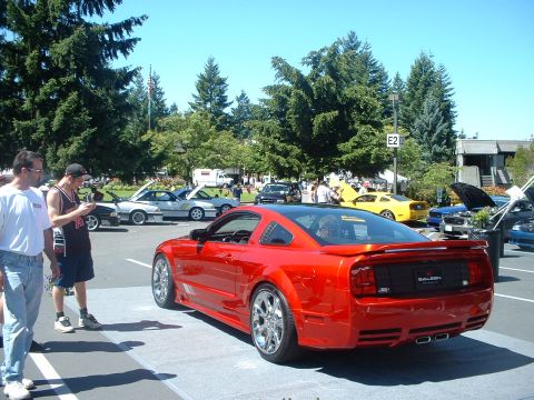 1999 Ford Crown Victoria - A red Saleen Mustang is displayed at an outdoor car show with other vehicles in the background.
