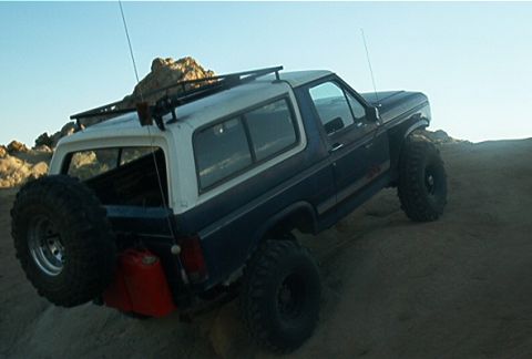 1986 Ford Bronco - 1986 Ford Bronco with external fuel cans and spare tire on rear, driving on dirt terrain.