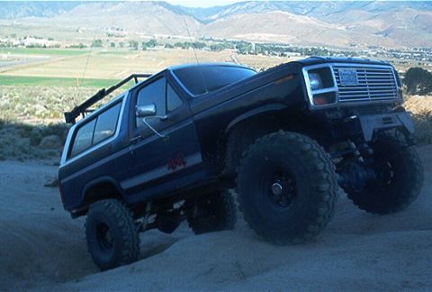 1986 Ford Bronco - A lifted Ford Bronco navigates a sandy incline with mountains in the background.