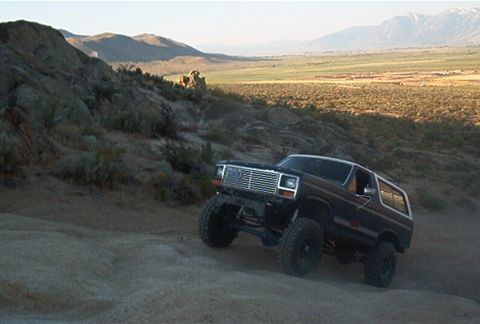 1986 Ford Bronco - A dark blue 1986 Ford Bronco with lifted suspension navigates a dirt road in a mountainous, arid landscape.
