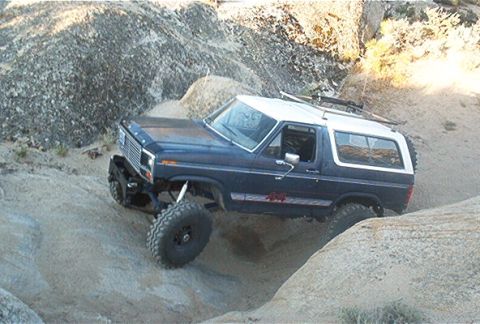 1986 Ford Bronco - This lifted Ford Bronco is shown navigating a challenging off-road course with large rocks and sandy terrain.