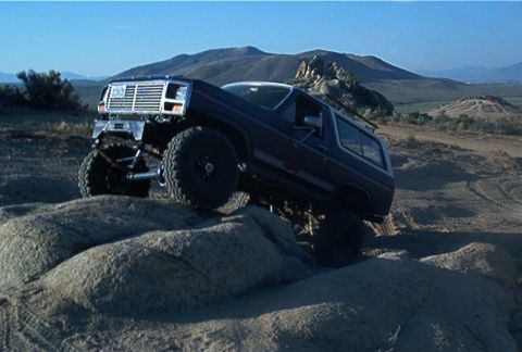 1986 Ford Bronco - A modified Ford Bronco navigates a rocky, off-road terrain under a clear blue sky.