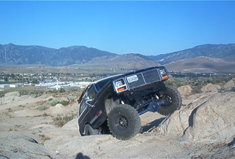 1986 Ford Bronco - This modified SUV is navigating a rocky incline under a clear blue sky.