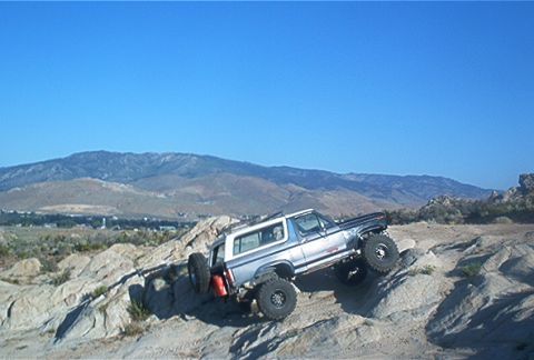 1986 Ford Bronco - A silver Ford Bronco navigates a rocky terrain under a clear blue sky.