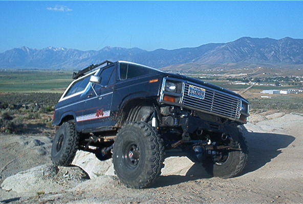 Modified SUV with large tires on rocky terrain, mountains in background. License plate reads BIGRNCH
