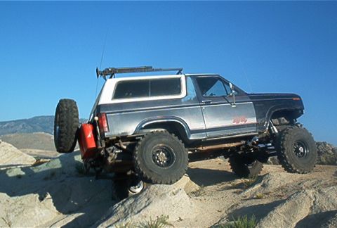 1986 Ford Bronco - Modified 1986 Ford Bronco with oversized tires and external fuel can navigates rocky terrain under a clear blue sky.