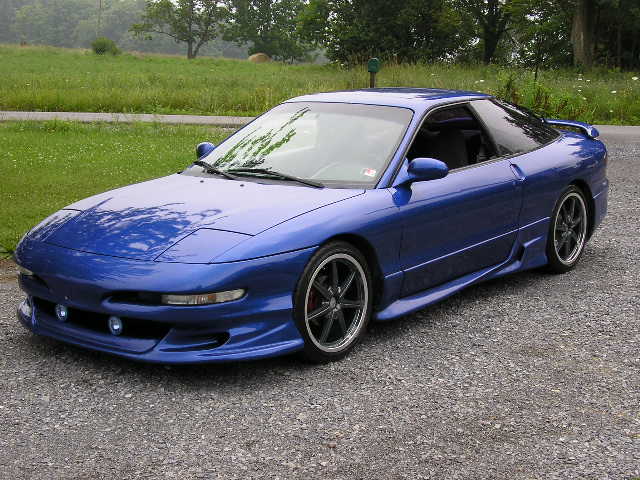 Blue 1994 Ford Probe parked on gravel lot with grassy field behind it