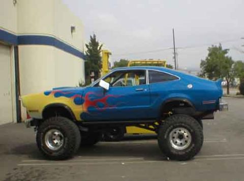 1990 Ford Bronco - blue compact hatchback coupe with red flame graphics sits jacked up on a lifted 4x4 truck chassis and off-road tires.