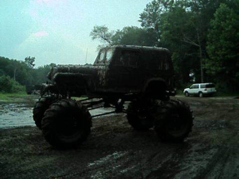 1987 Suzuki Samurai - A heavily modified off-road vehicle, covered in mud, sits in a muddy clearing with a white SUV visible in the distance.