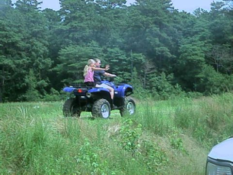 1987 Suzuki Samurai - Two people ride a blue ATV through a grassy field with a wooded area behind them.
