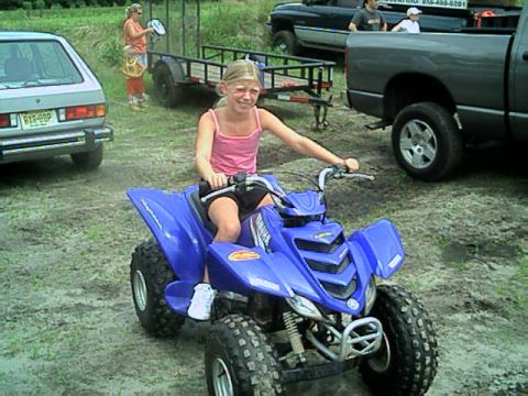 1987 Suzuki Samurai - A young girl rides a blue ATV in a dirt lot, with other vehicles and a trailer visible in the background.