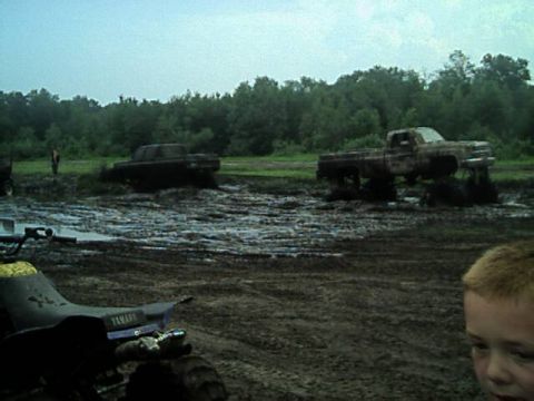 1987 Suzuki Samurai - Two trucks are deeply stuck in a muddy field with trees in the background.
