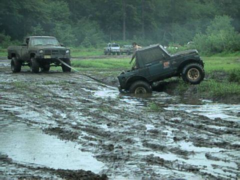 1987 Suzuki Samurai - Two off-road vehicles struggle in deep mud, with one truck attempting to tow the other out of a mire.