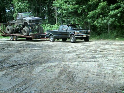 1987 Suzuki Samurai - A black pickup truck tows a trailer carrying a heavily modified monster truck through a wooded area.