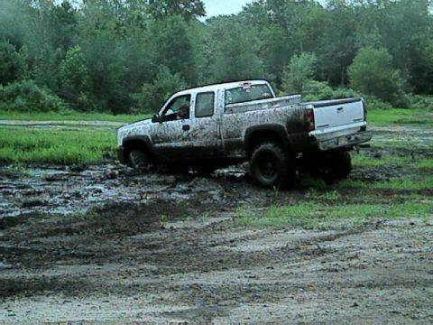 1987 Suzuki Samurai - A white pickup truck is covered in mud as it navigates through a muddy field with trees in the distance.