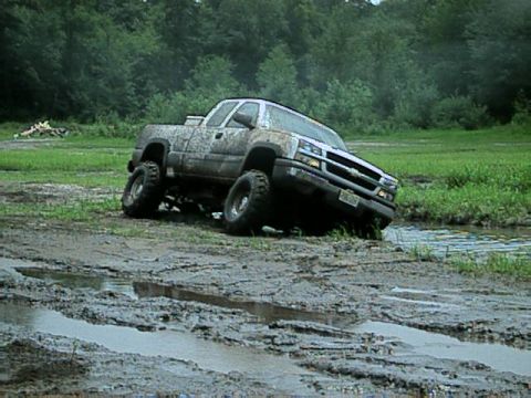 1987 Suzuki Samurai - A lifted pickup truck navigates a muddy off-road course with trees and water visible.