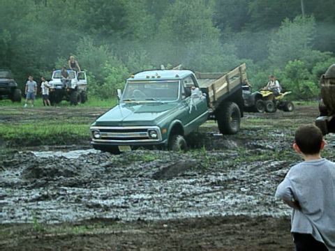 1987 Suzuki Samurai - A group of people watch as a green pickup truck gets stuck in a muddy area, with other vehicles visible in the background.