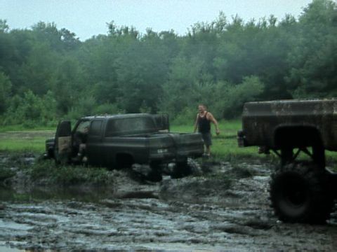 1987 Suzuki Samurai - Two trucks are stuck in a muddy field, with a person standing between them and trees in the background.