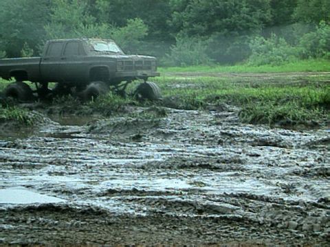1987 Suzuki Samurai - A heavily modified pickup truck is stuck deep in the mud, surrounded by overgrown grass and trees.