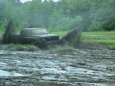 1987 Suzuki Samurai - A dark pickup truck is stuck in a muddy field, with black mud spraying from its rear wheels.