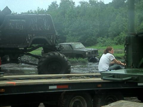 1987 Suzuki Samurai - Several muddy off-road vehicles are stuck in a muddy field, with a woman sitting on a trailer in the foreground.