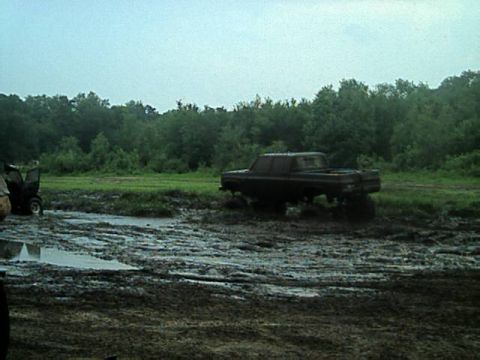 1987 Suzuki Samurai - A dark pickup truck is stuck in a muddy field with trees in the distance. Another vehicle is partially visible on the left.