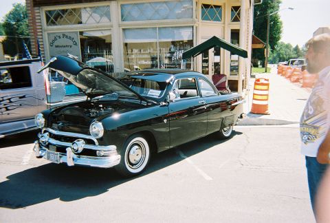 1972 Ford Ranchero - A classic black coupe from the 1950s is displayed with its hood up, parked next to a building.