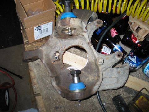 1995 Ford Bronco - Rusty cast steering knuckle laid on a wood plank in a garage with brand-new upper and lower ball joints installed, their blue polyurethane…