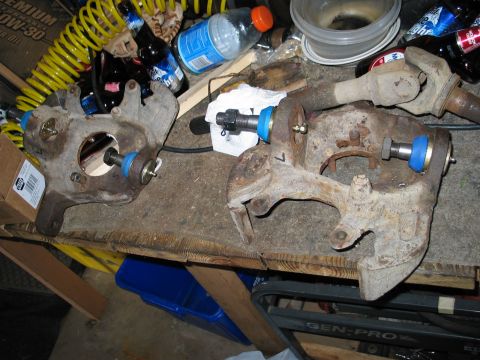 1995 Ford Bronco - Two rusty steering knuckles laid on a carpeted workbench mid-rebuild, each fitted with a new blue-collared ball joint and a threaded press…