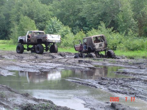 1995 Dodge 1500 - Two lifted off-road trucks are stuck in a muddy terrain with trees in the background.