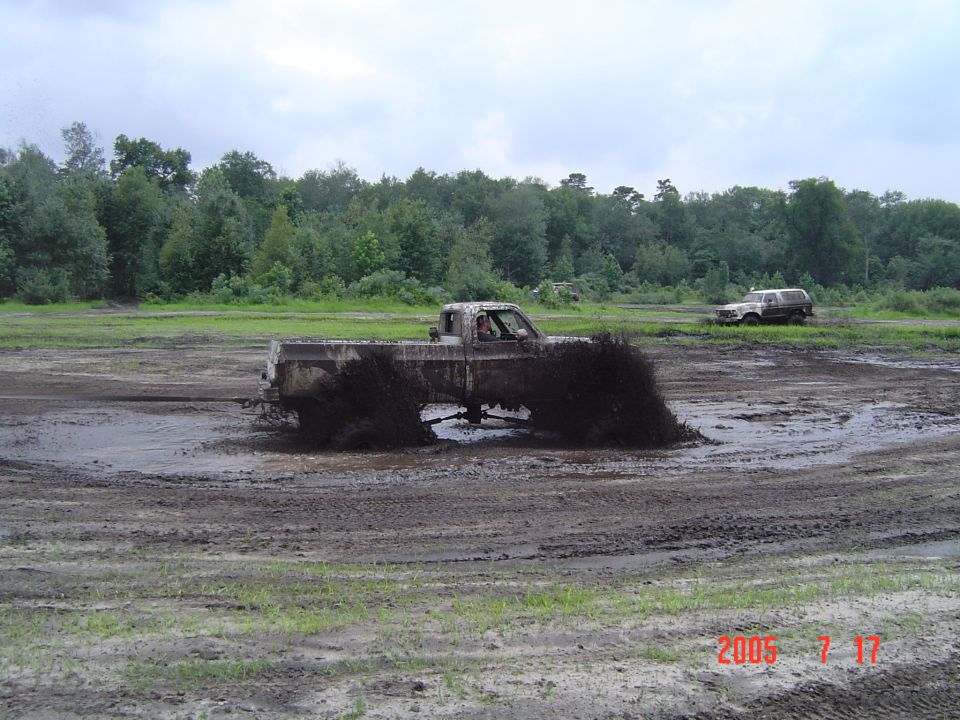 Mud-splattered pickup truck driving through a muddy field, side view. Another SUV is visible in the background