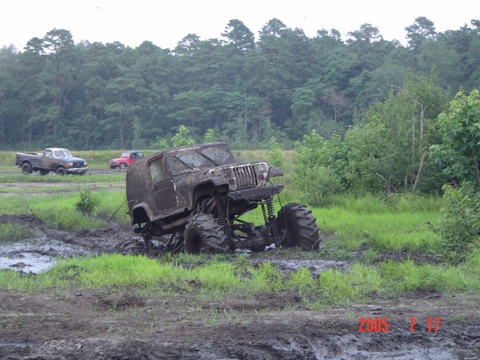 Mud-covered Jeep Wrangler with oversized tires stuck in mud, two trucks in background