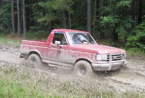 1988 Ford Bronco - A red pickup truck navigates a muddy trail surrounded by trees.