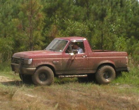 1988 Ford Bronco - A muddy red SUV drives through a grassy, wooded area.