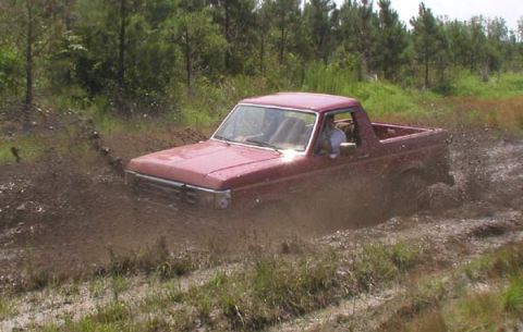 1988 Ford Bronco - A red pickup truck navigates a muddy trail, kicking up dirt and water as it goes.