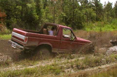 1988 Ford Bronco - Red Ford Bronco navigating a muddy trail with trees in the background.