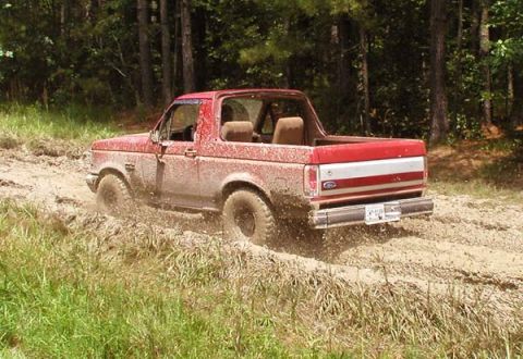 1988 Ford Bronco - A red SUV navigates a muddy trail surrounded by trees.