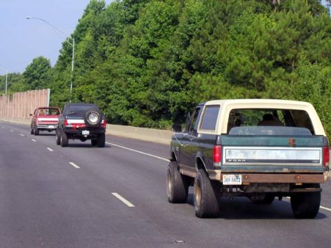 1988 Ford Bronco - A dark green and tan Ford Bronco is seen from behind on a highway, with another vehicle following.