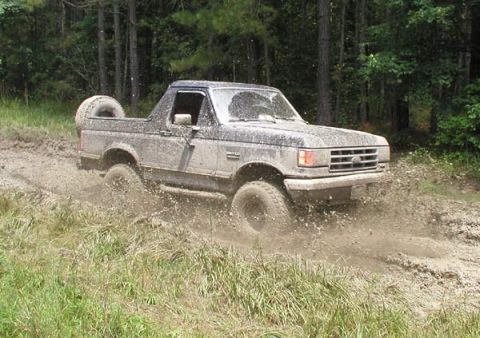 1988 Ford Bronco - A muddy Ford Bronco navigates a wet, wooded trail, sending dirt flying.