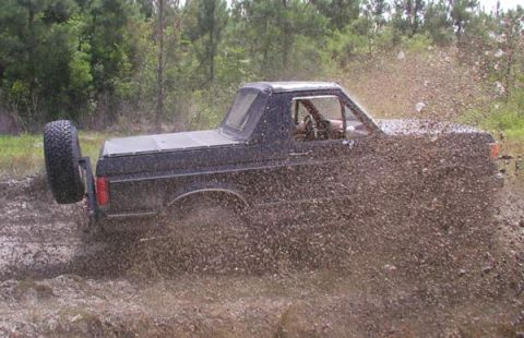 1988 Ford Bronco - A black Ford Bronco churns up mud as it drives through a muddy terrain.