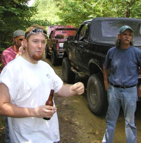 1988 Ford Bronco - Several off-road vehicles are parked on a dirt path in a wooded area with people standing around.