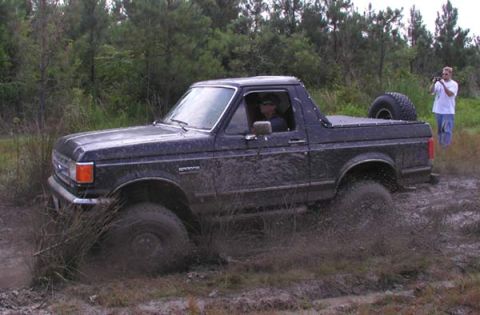 1988 Ford Bronco - A dark blue Ford Bronco navigates a muddy off-road course with a person filming in the background.