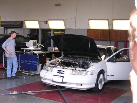 1993 Ford Taurus - A white sedan is on a dynamometer in a garage, with its hood open and a person standing next to it.