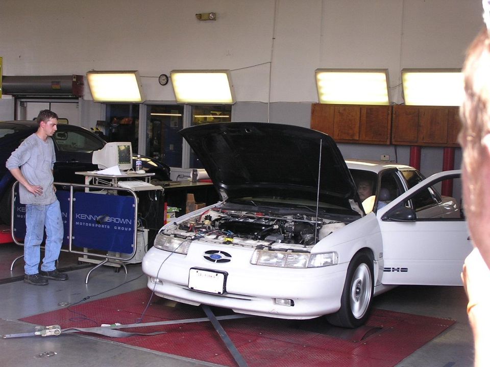 white sedan on a dynamometer with hood open, guy in jeans standing nearby