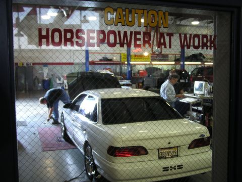 1993 Ford Taurus - A white sedan is on a dynamometer in a garage, with a sign that reads 'CAUTION HORSEPOWER AT WORK'.