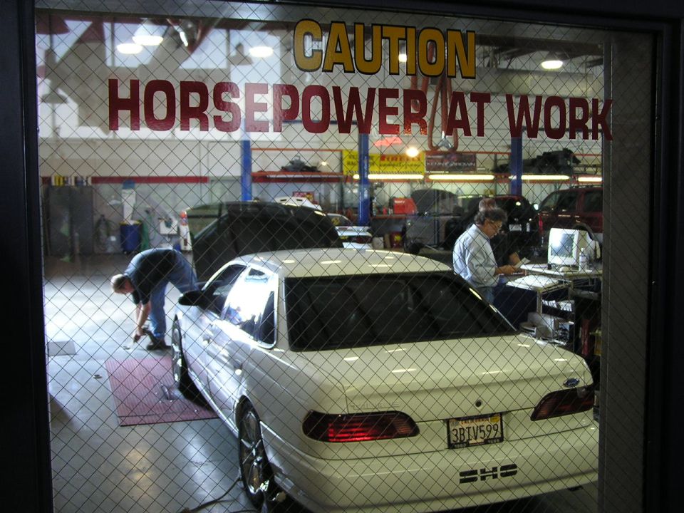 White sedan with a license plate reading 3BIV599, viewed from the rear in a garage with 'CAUTION HORSEPOWER AT WORK' sign